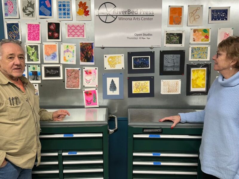 A man in a brown shirt and woman in a light blue sweater stand in front of dark green cabinets and small pieces of art.