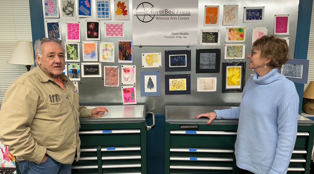 A man in a brown shirt and woman in a light blue sweater stand in front of dark green cabinets and small pieces of art.