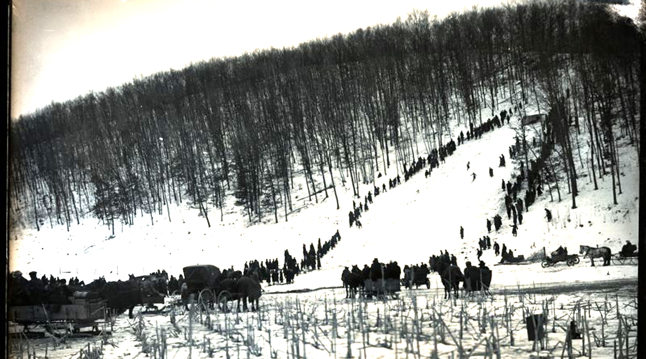 Black and white photo of a ski hill with hundreds of standing trees and people standing along the downhill ski trail.