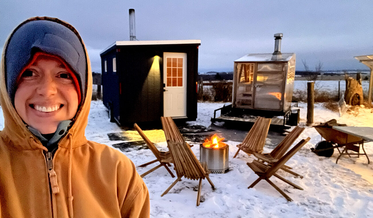 A woman bundled up in a brown jacket with gray hoodie smiles in the forefront of the picture. Behind her is a circle of wooden chairs around a metal fire pit and two portable saunas.