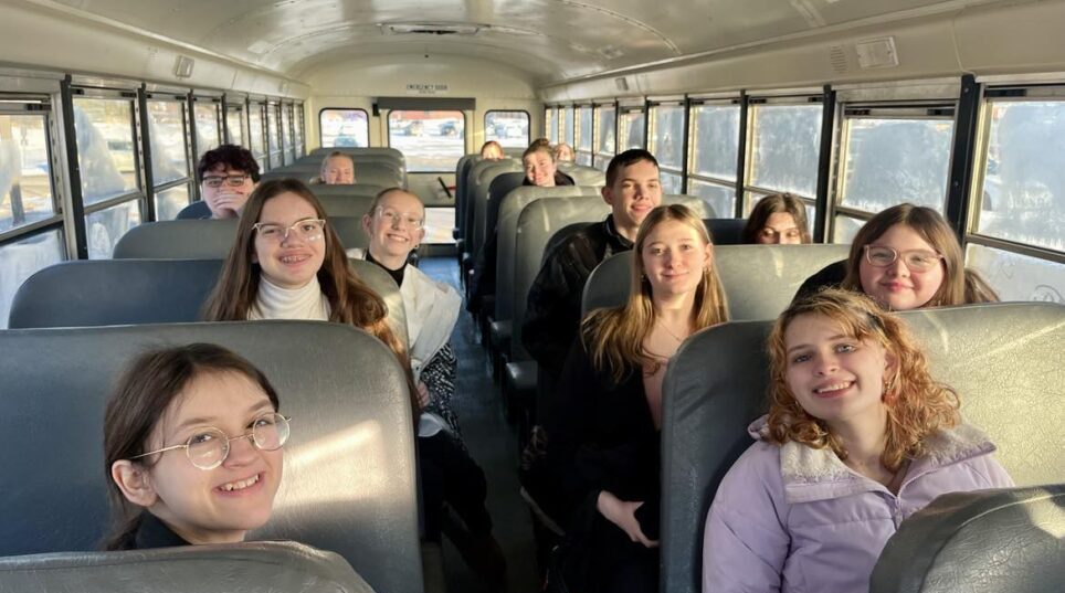 Teenage students sit in gray bus seats and look at the camera with smiles.