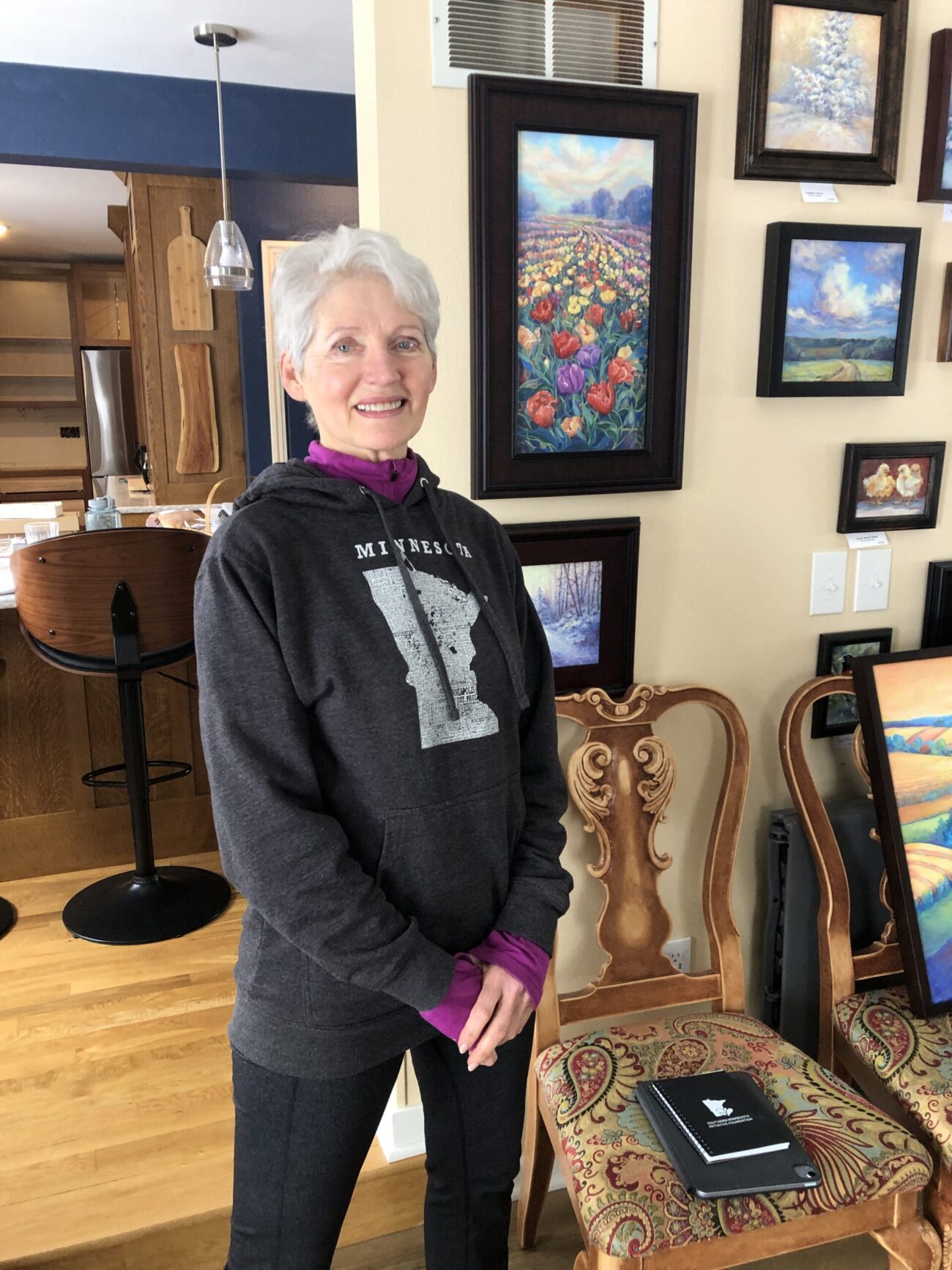 A woman with gray hair wearing a black sweatshirt with the silhouette of Minnesota in white, stand by an upholstered chair in front of colorful landscape oil paintings.