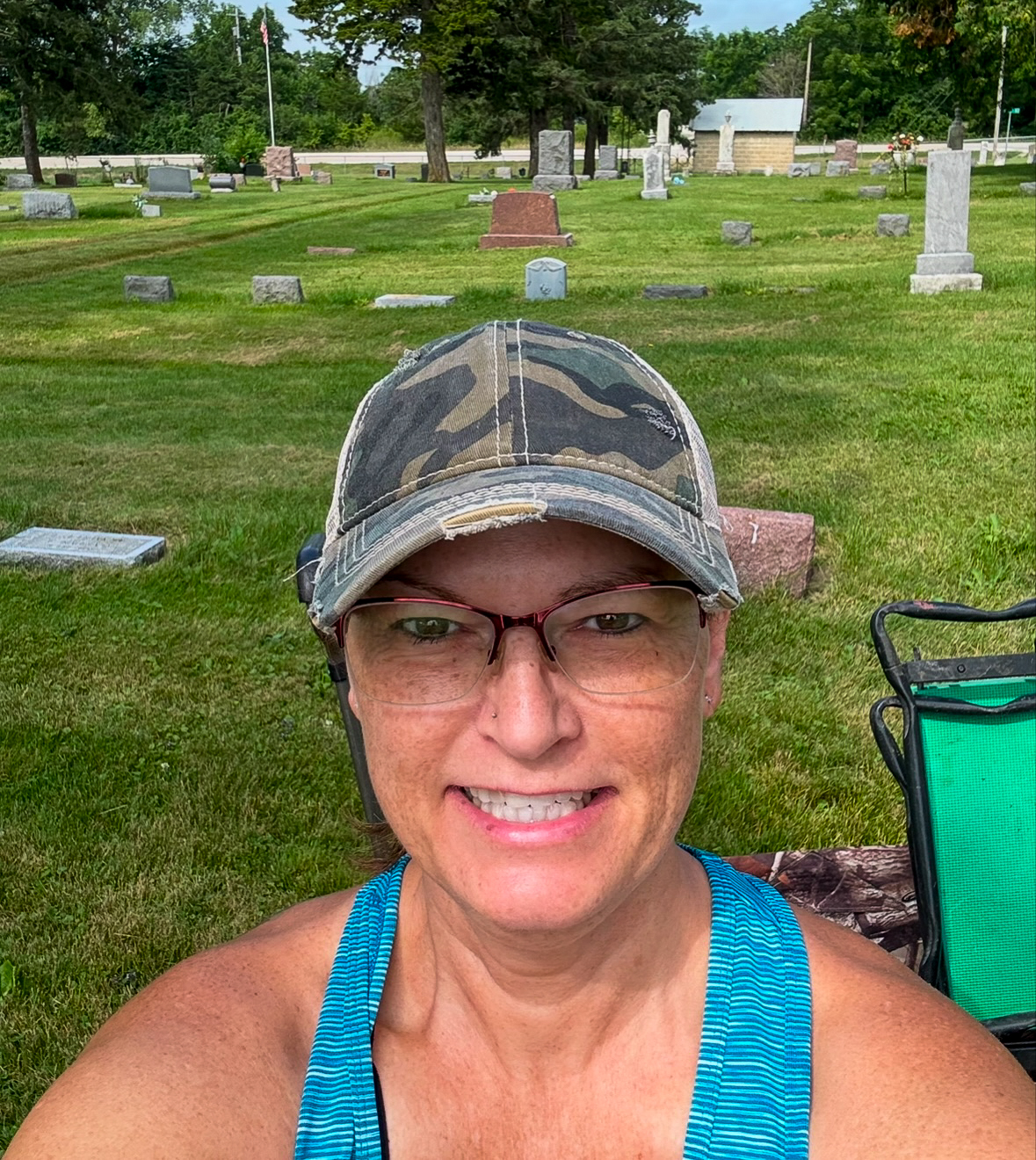 Woman looks up at the camera with a grassy cemetery behind her. She is wearing a gray ball cap and eyeglasses and a teal green tank top.