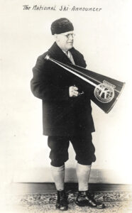 A black and white photo of a man wearing knee length dark pants, a long sleeved dark jacket, and a dark beanie type hat. He is holding a large megaphone with an emblem on the side.