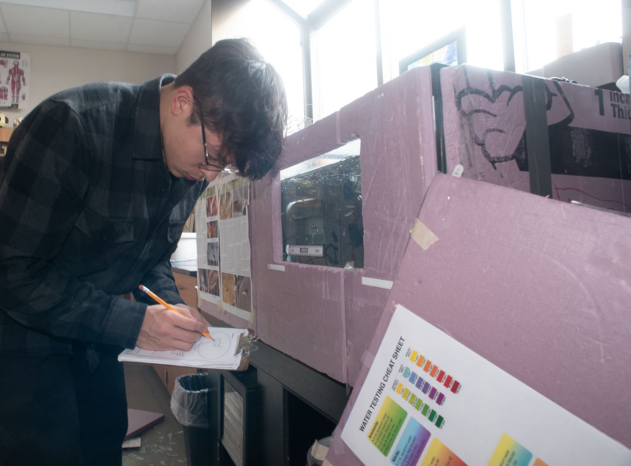 A high school boy with a long sleeve shirt, black hair, and glasses holds a clipboard and records a number with a pencil.