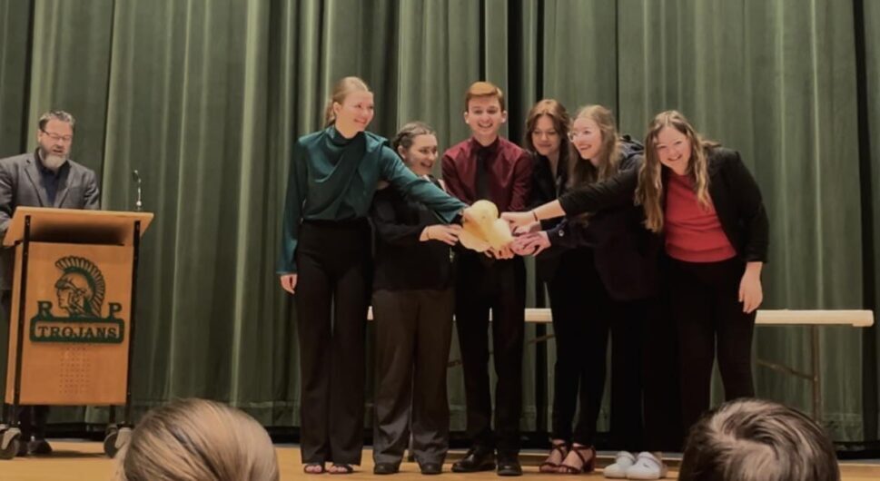 A group of teenage students stand on a stage in front of a green curtain. They all reach to the middle to hold a yellow stuffed duck.