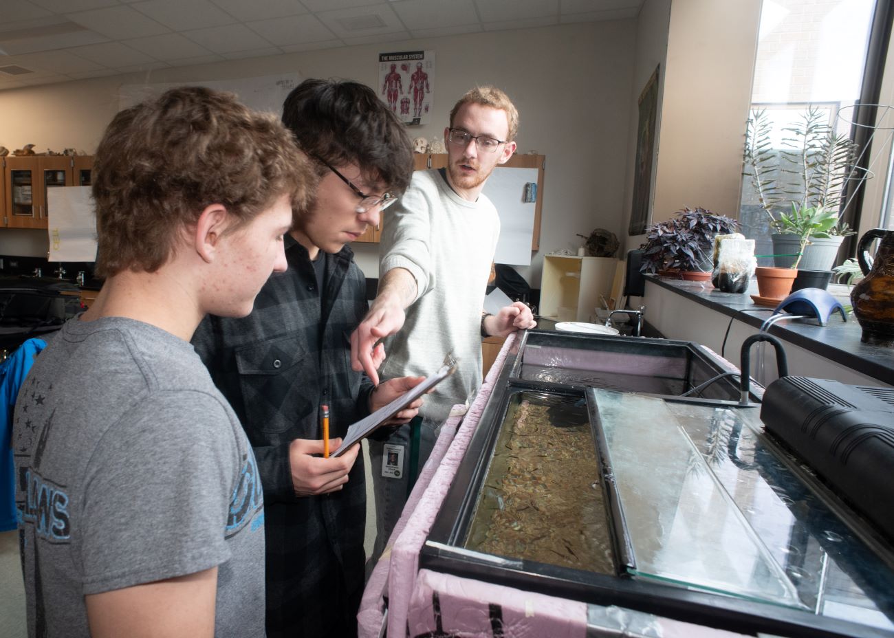 Two high school boys stand in front of a large open fish tank while the older man explains how to measure the water.