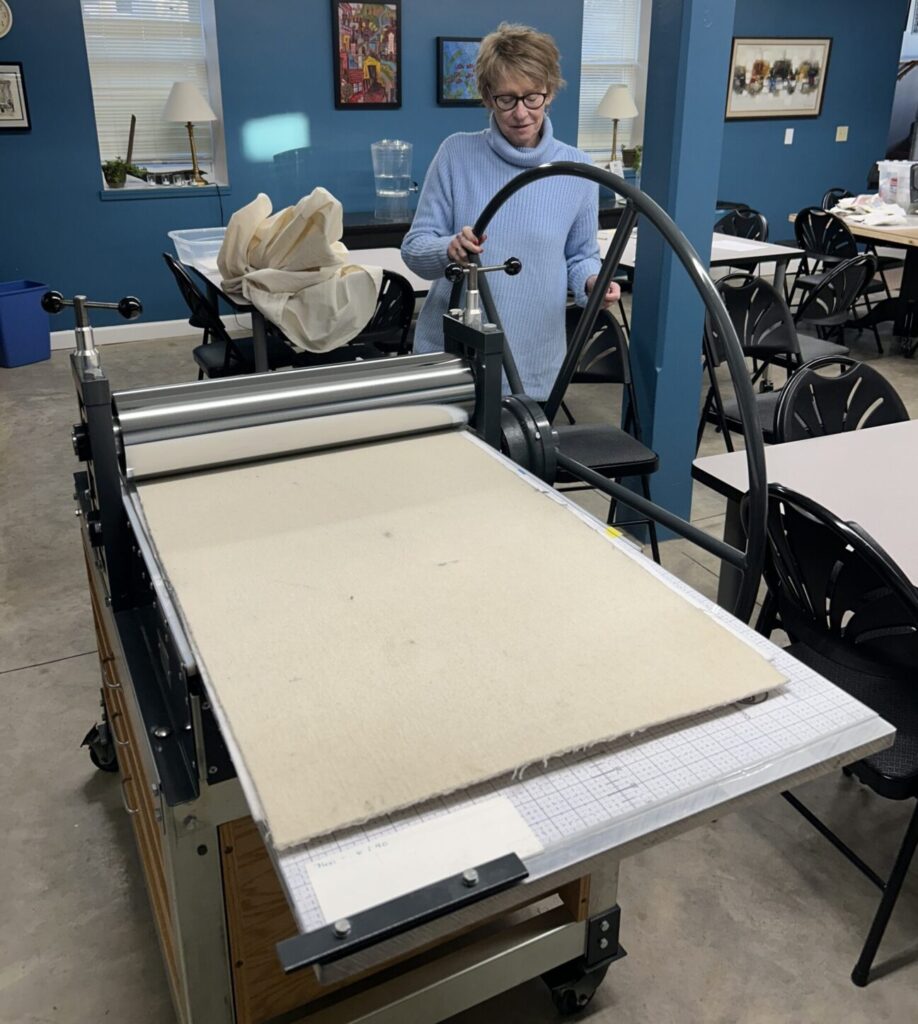 Woman in light blue sweater stands in front of a large metal wheel attached to an long table printing press.