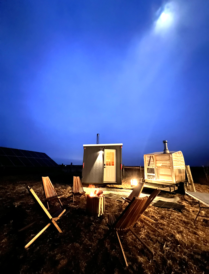 A dark blue sky with moonlight shining through clouds lights up two portable saunas glowing and lit from the inside.