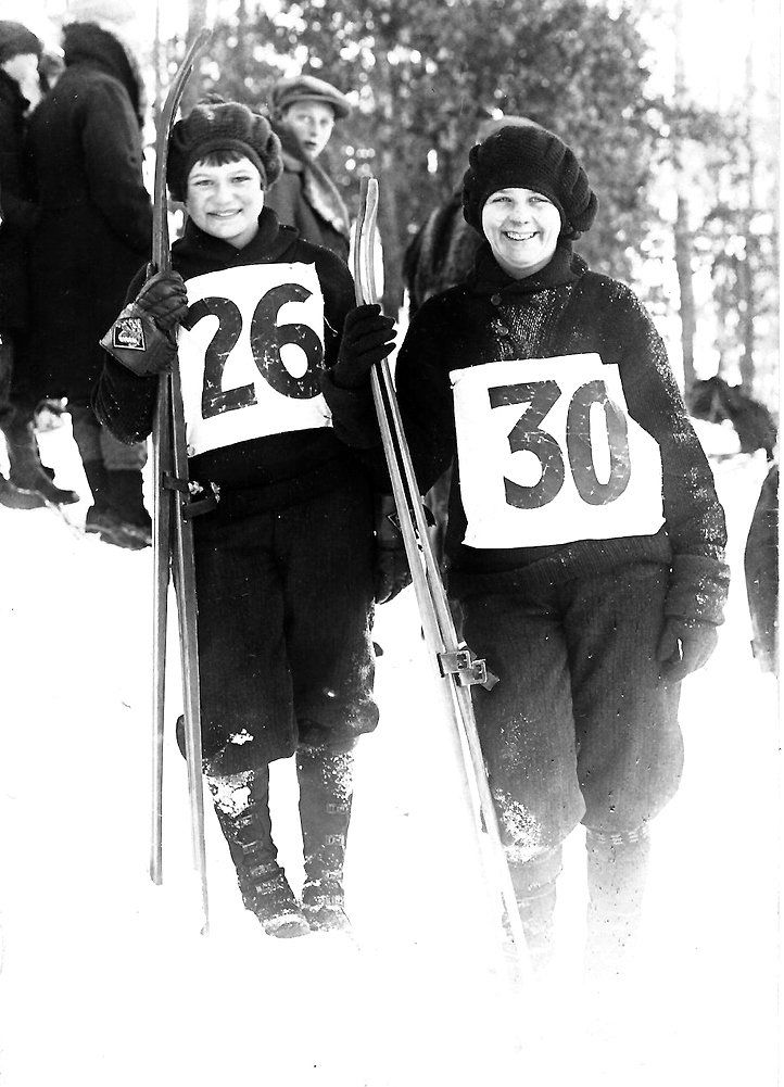 Black and white photo of two female skiers outside wearing dark winter clothes and hats with large white labels on the fronts of their shirts with the numbers 26 (on the left) and 30 (on the right).