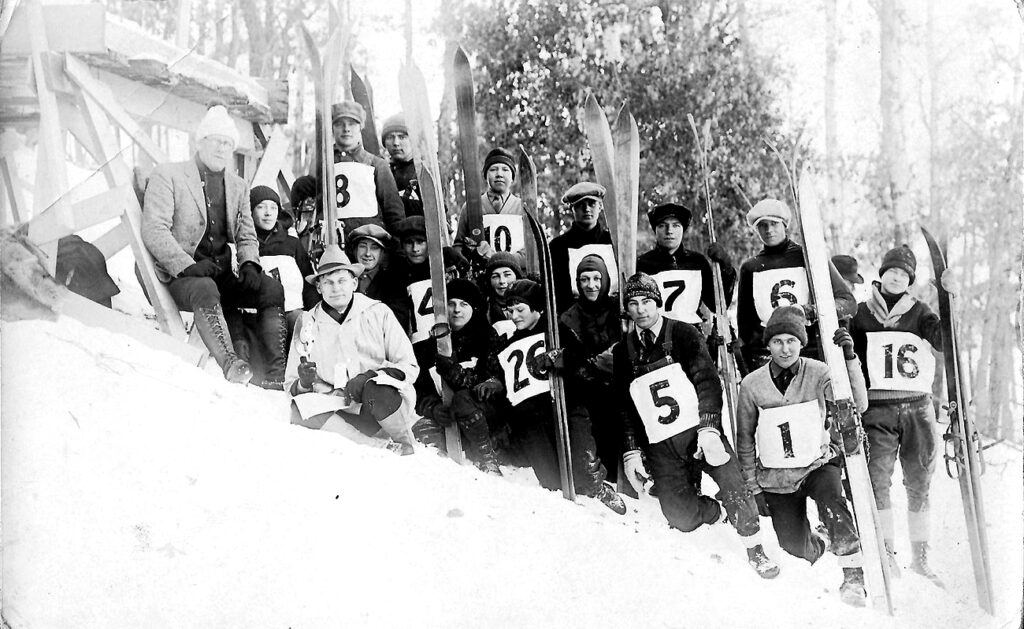 Black and white photo of a group of skiers standing on an inclined hill with dark clothing and large white labels on the fronts with numbers.