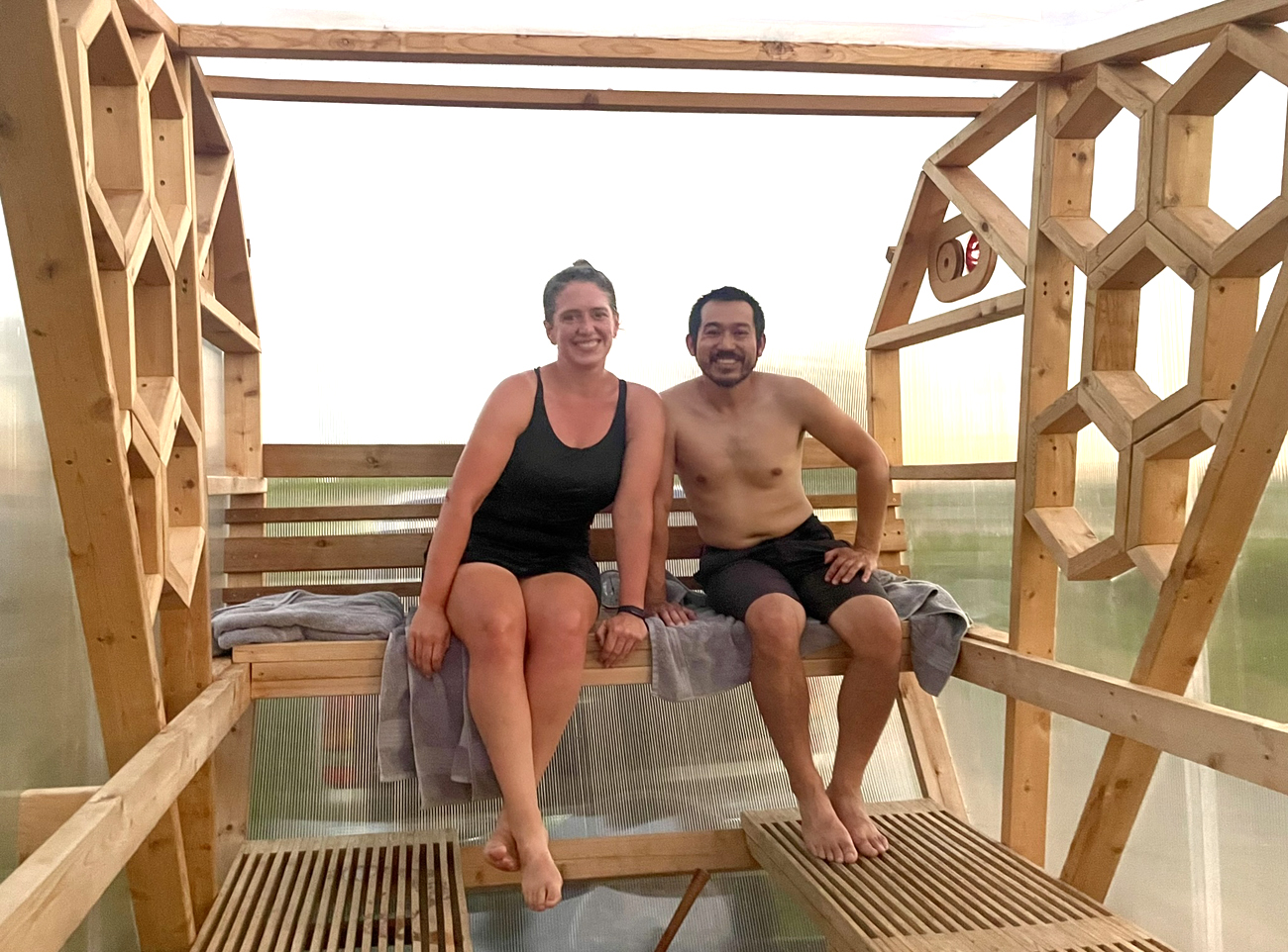 A woman in a black tank top and a man with no shirt and black shorts sit on a bench in a see through sauna made from wood and plastic corrugated panels.