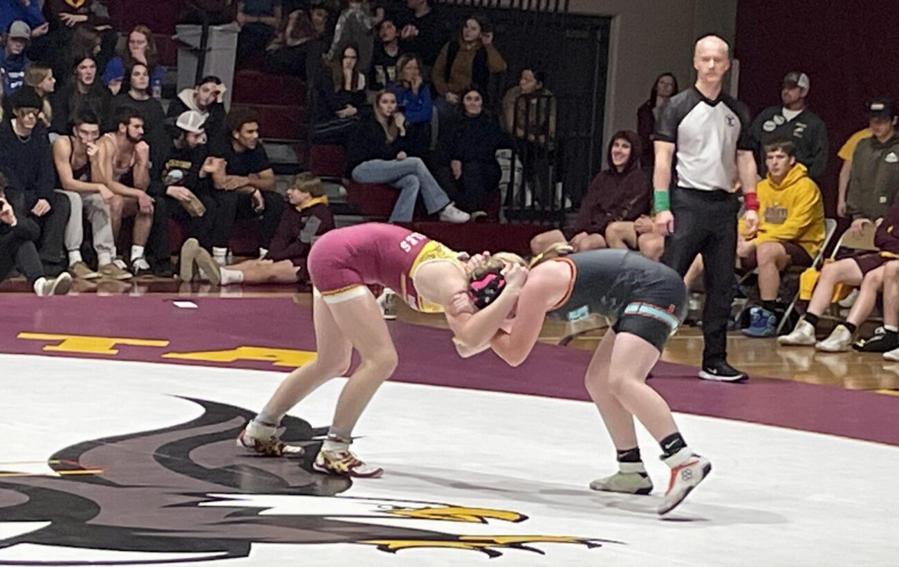 A wrestler in a maroon singlet ties up at the head and shoulders with a wrestler in a black singlet. They are wrestling on a while mat with an emblem of a brown hawk. A referee watches from the sidelines.