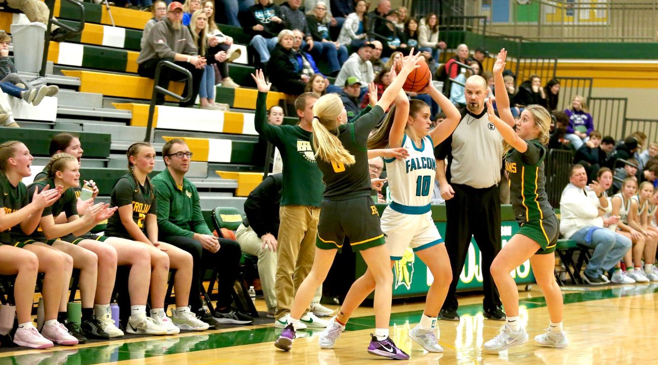 Girl in white basketball uniform with green trim and numbers holds the basketball over her head while her opponent in black tries to steal the ball. A referee with whistle in his mouth watches.