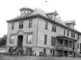 Black and white photo of an old two-story rectangular building with windows wrapping around the building.