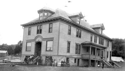 Black and white photo of an old two-story rectangular building with windows wrapping around the building.