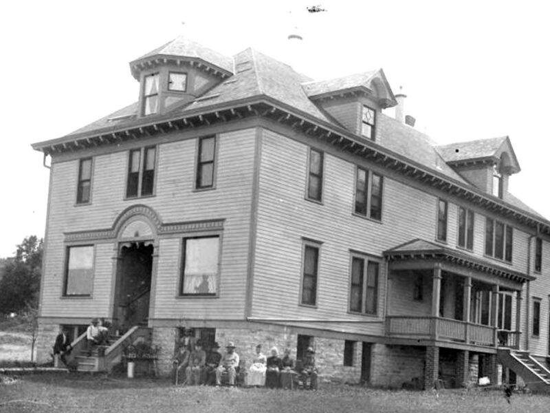 Black and white photo of an old two-story rectangular building with windows wrapping around the building.