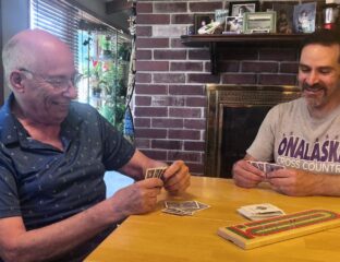 Two men, one older and one younger, play cribbage on a wood table in front of a red brick fireplace.