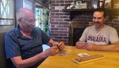 Two men, one older and one younger, play cribbage on a wood table in front of a red brick fireplace.