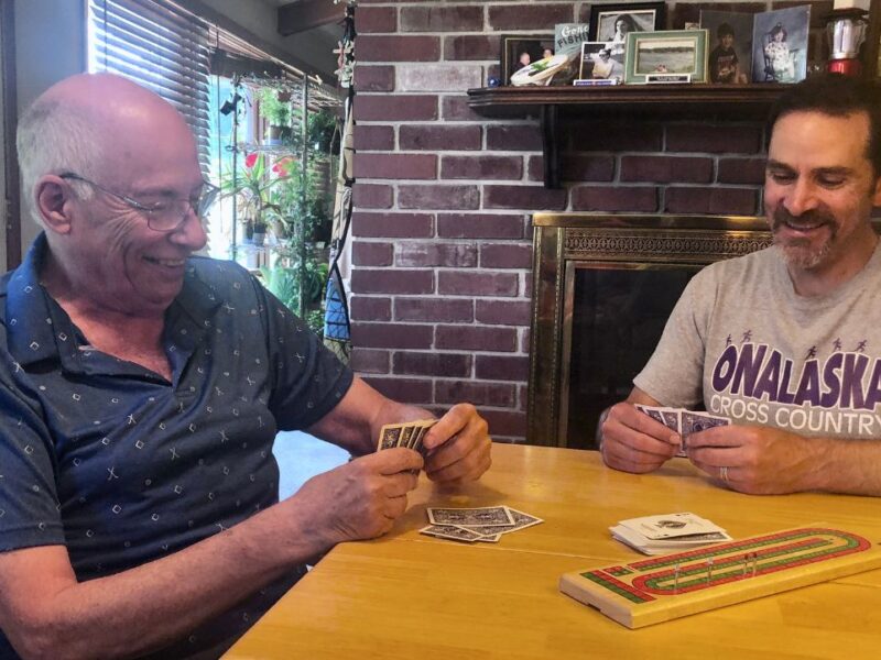 Two men, one older and one younger, play cribbage on a wood table in front of a red brick fireplace.