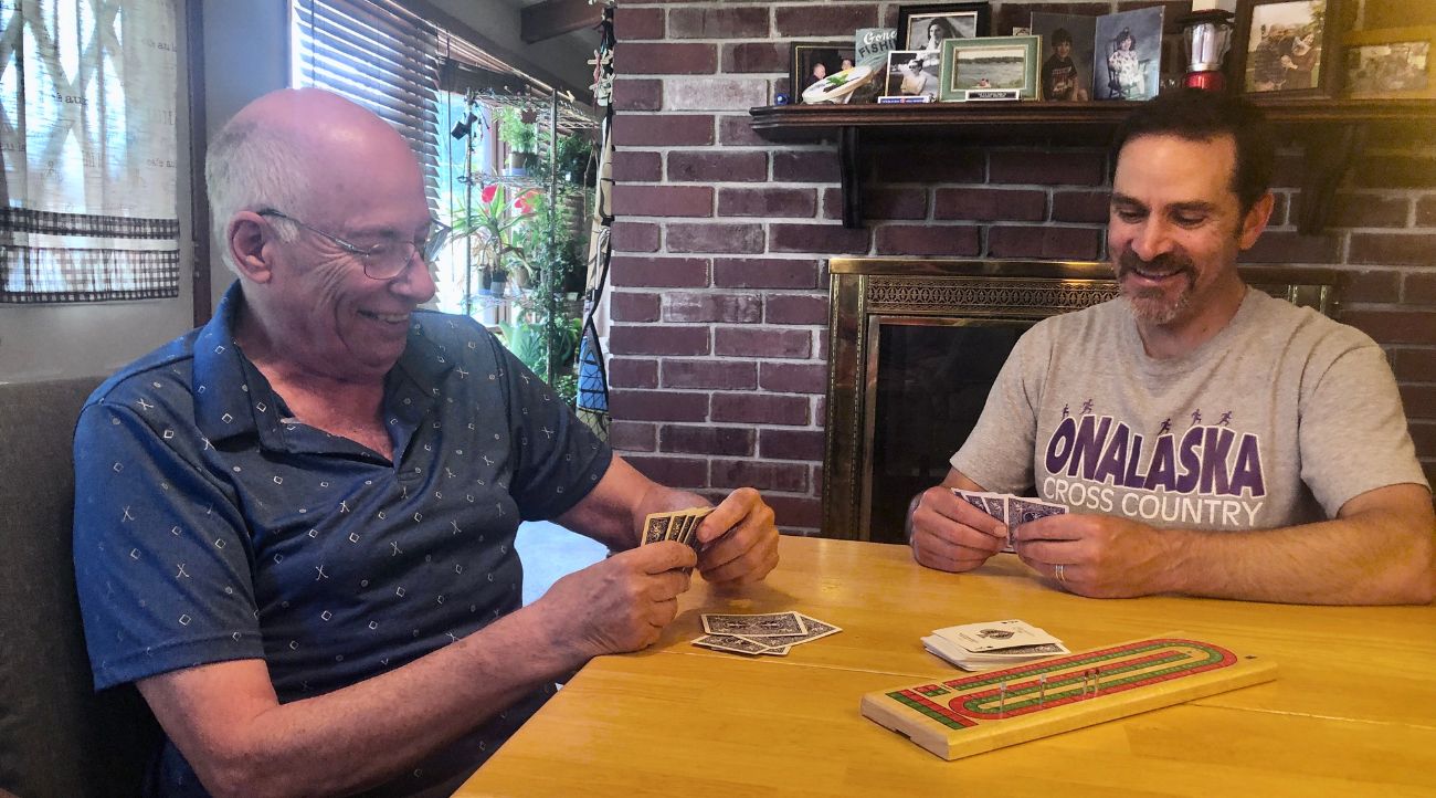 Two men, one older and one younger, play cribbage on a wood table in front of a red brick fireplace.