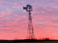 An old fashioned metal windmill stands above a field against a sky of pink and blue clouds at sunset