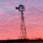 An old fashioned metal windmill stands above a field against a sky of pink and blue clouds at sunset