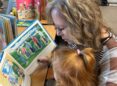 A woman with blonde-brown hair reads a picture book to a child with red hair pulled back in a partial pony tail.