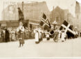 A black and white photo of women wearing long skirts marching in a parade under various Scandinavian flags.