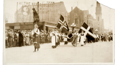 A black and white photo of women wearing long skirts marching in a parade under various Scandinavian flags.