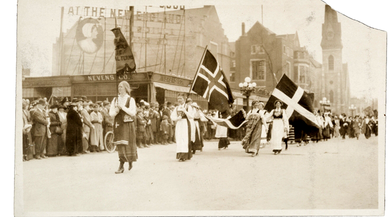 A black and white photo of women wearing long skirts marching in a parade under various Scandinavian flags.