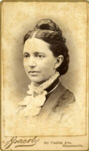 A black and white photo of a women wearing a frilly white collar with a bow, her hair is parted down the middle and wrapped on top of her head in a bun.