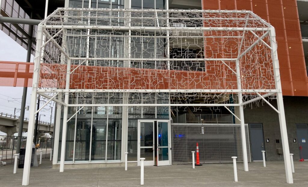 An open metal sculpture shaped like a house with a lattice like roof and open space underneath. Sculpture sits next to a large red building with large windows trimmed in silver metal.