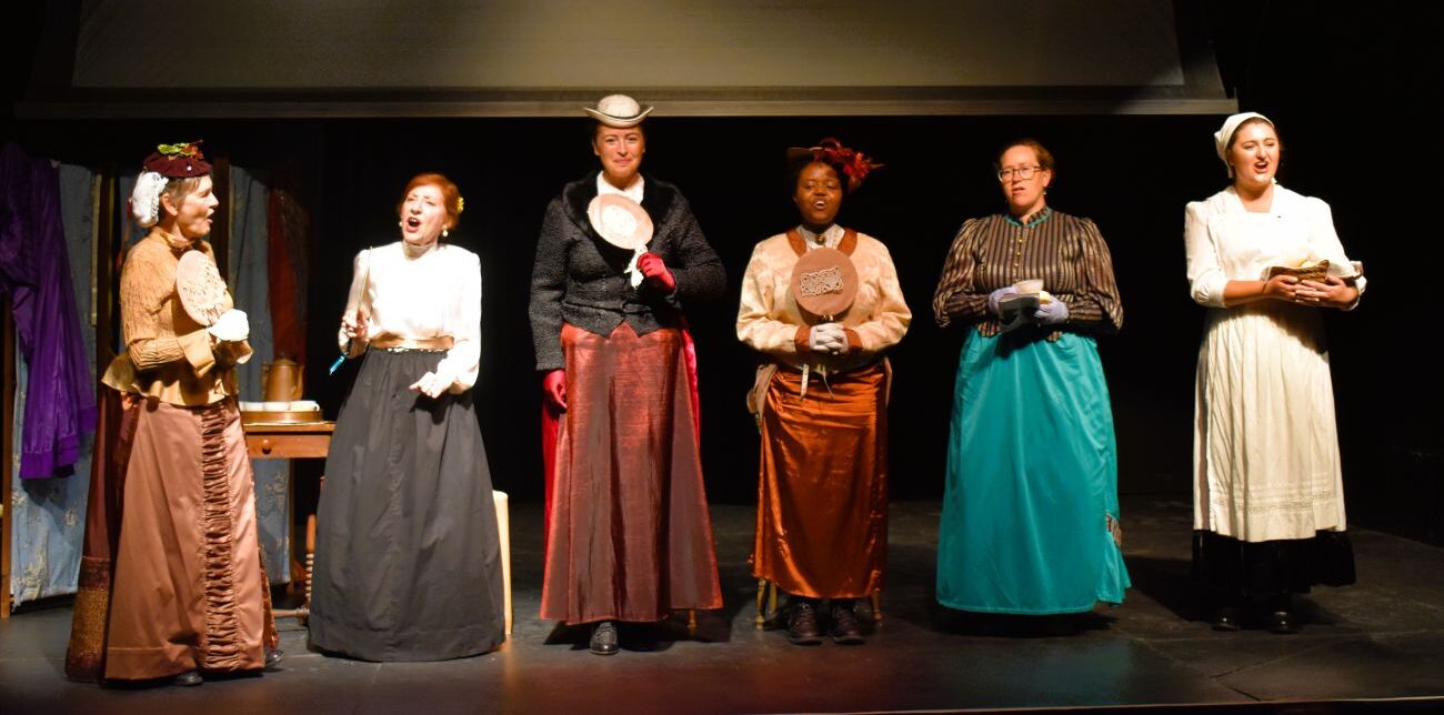 Six woman wearing historical clothing perform on a stage.