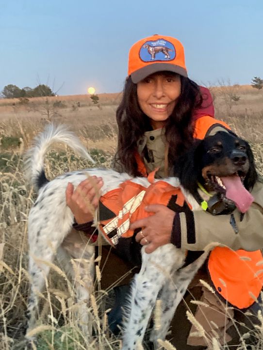 Women with orange cap and hunting vest holds an English Setter dog who's panting. Behind her is a brown field with a full moon just peeking over the horizon.