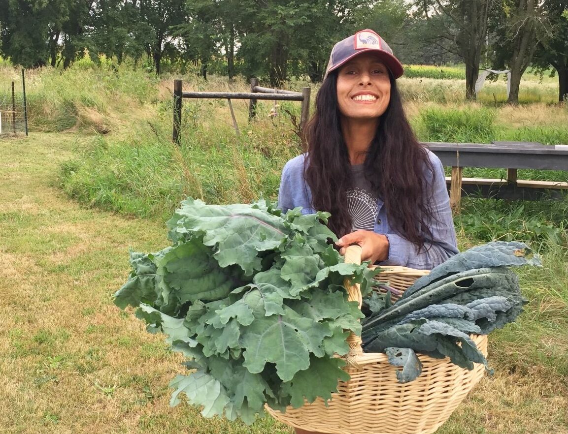 Woman with long black hair wearing a ballcap holds a large wicker basket filled with giant leaves of kale.