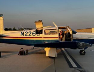 A small two-seater white and blue airplane sits on the tarmac with the door open and a women peeking out and smiling.