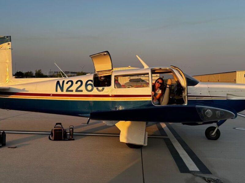A small two-seater white and blue airplane sits on the tarmac with the door open and a women peeking out and smiling.