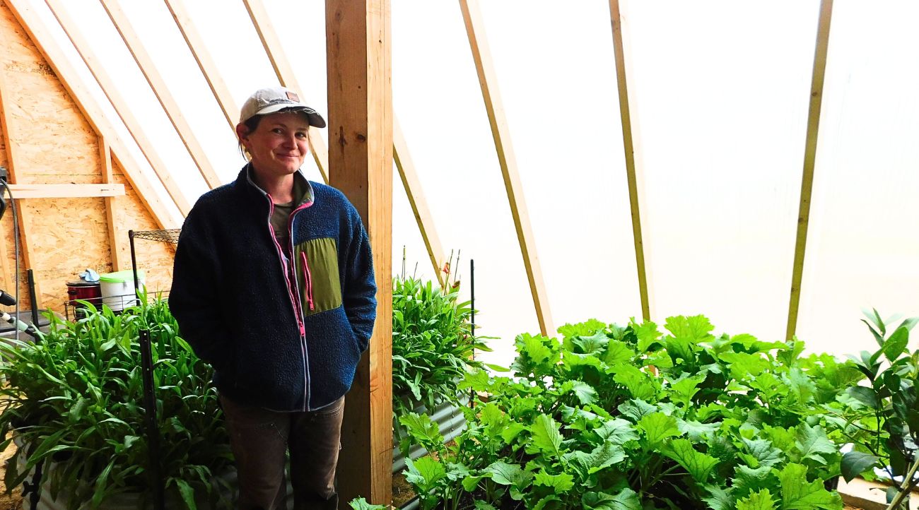 A woman in a ball cap and light jacket stands inside a greenhouse surrounded by green, leafy plants.