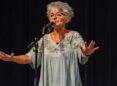An older woman with short gray hair stands behind a microphone on a stand and in front of a black curtain. Her arms are outstretched and her hands are flat with fingers spread widely.