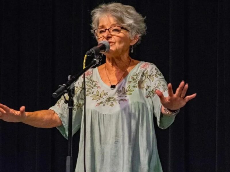 An older woman with short gray hair stands behind a microphone on a stand and in front of a black curtain. Her arms are outstretched and her hands are flat with fingers spread widely.