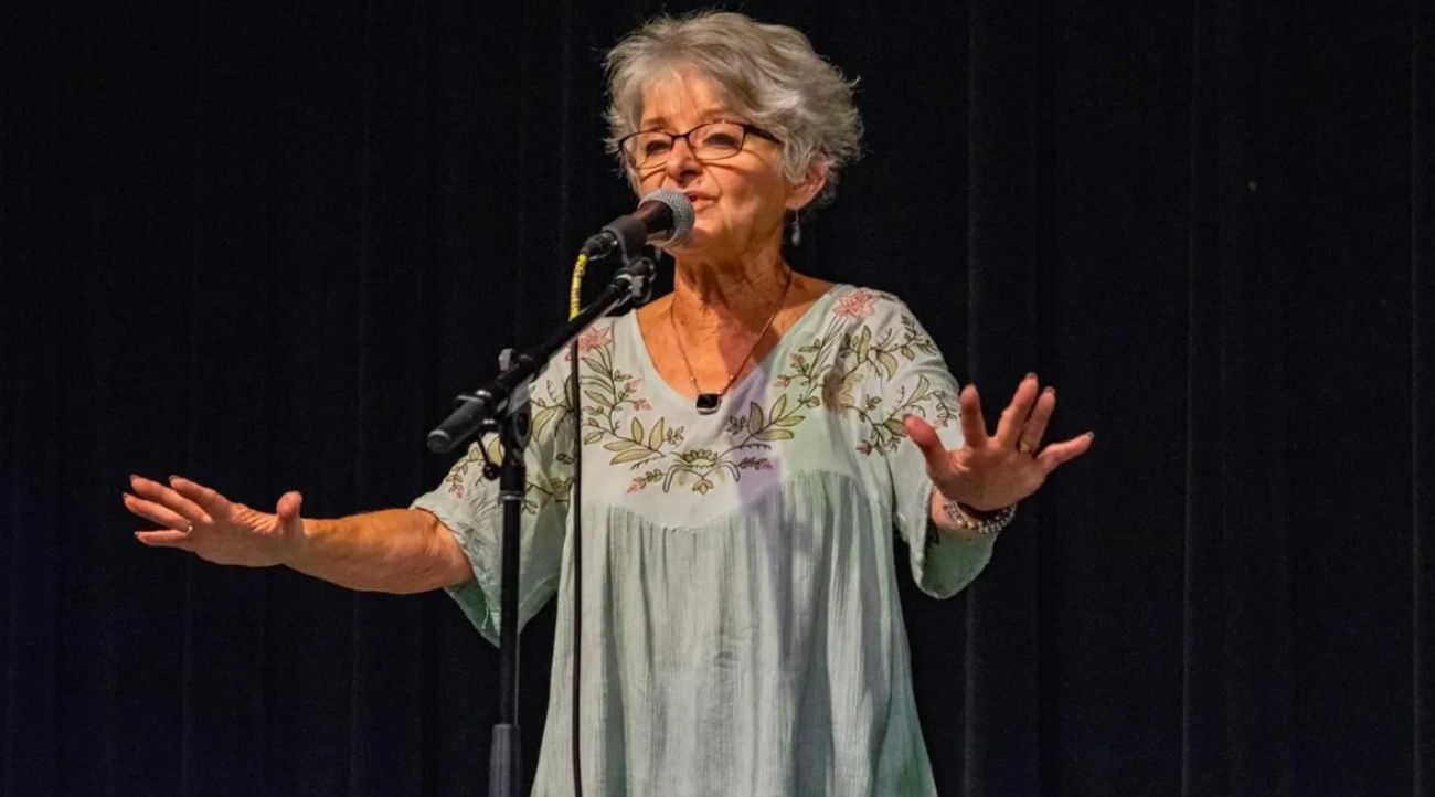 An older woman with short gray hair stands behind a microphone on a stand and in front of a black curtain. Her arms are outstretched and her hands are flat with fingers spread widely.