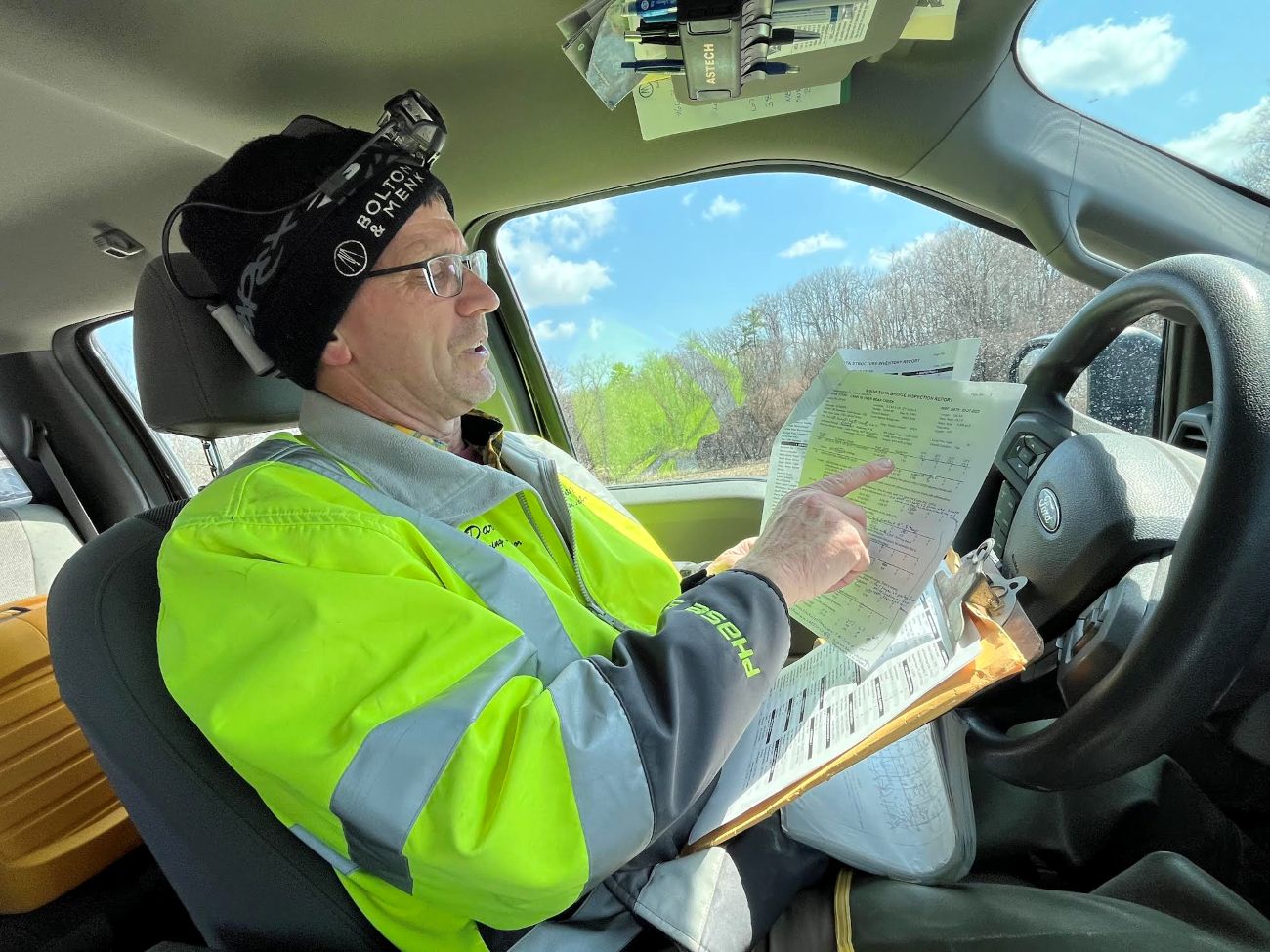 A man in a bright yellow vest sits in a pickup and points at a sheet of paper with rows of numbers.