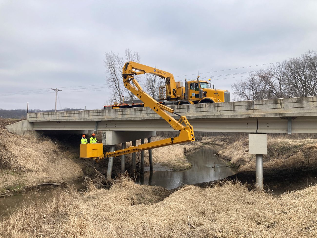 A yellow construction truck sits on top of a bridge. A long, folding arm extends out over the bridge with a bucket at the end holding two men hovers underneath the bridge.