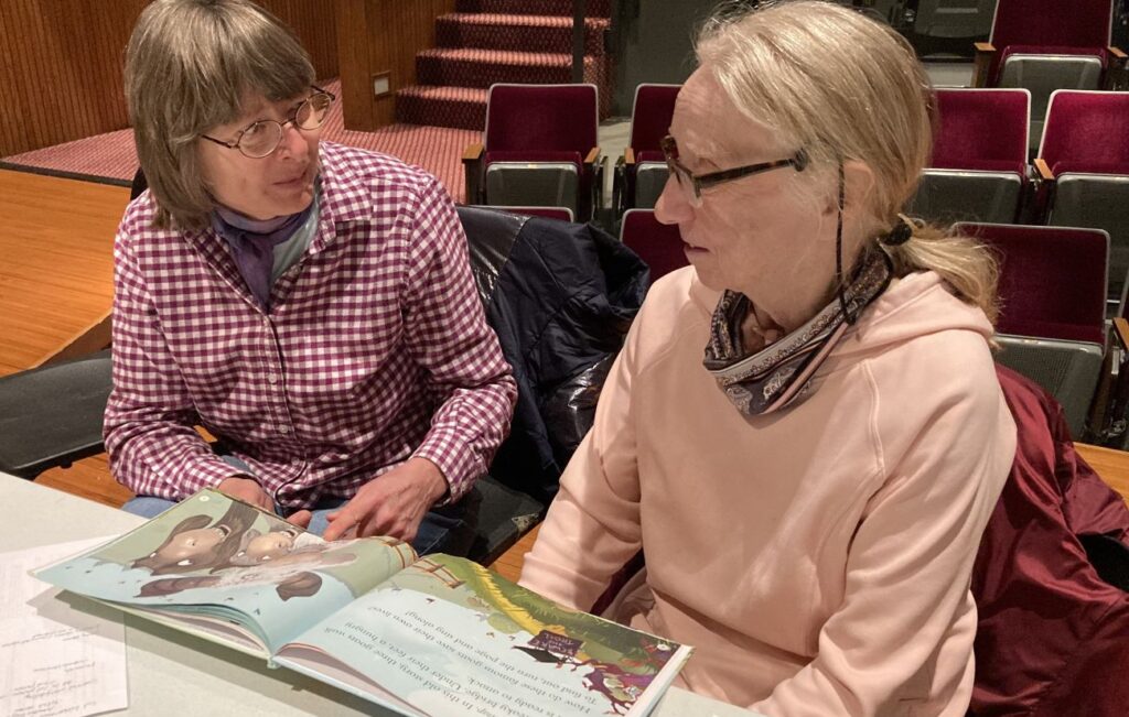Two women look at each other as they sit in front of a white plastic table with an illustrated children's book in from of them opened to a page with troll drawings.