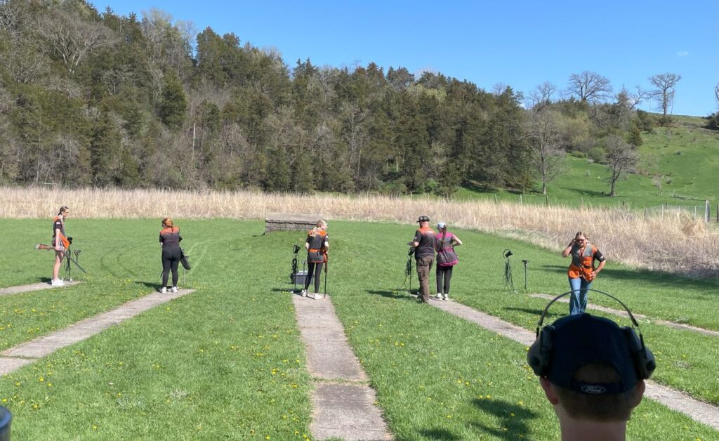 Students stand in a grassy field with long cement strips. They are holding and loading shotguns in preparation for trap shooting practice.