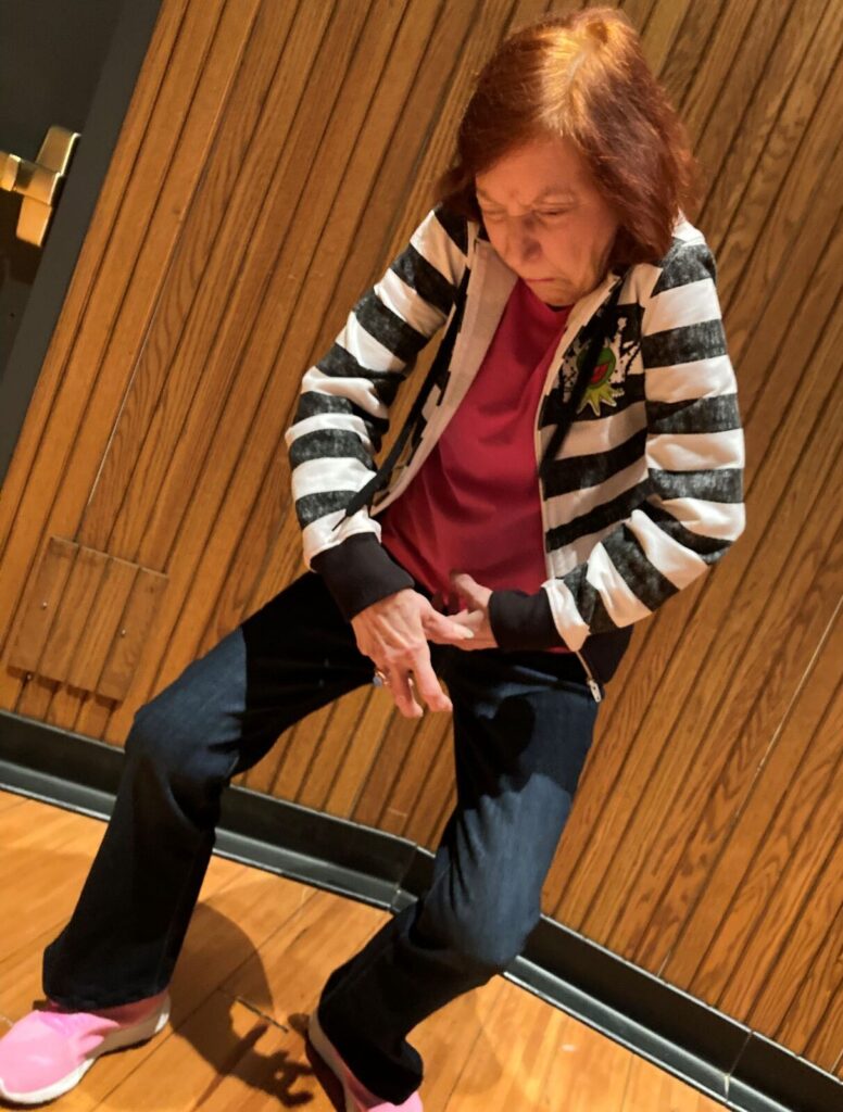 An older woman with red hair and wearing a black and white wide striped zipper sweatshirt crouches forward with her hand sin front of her.