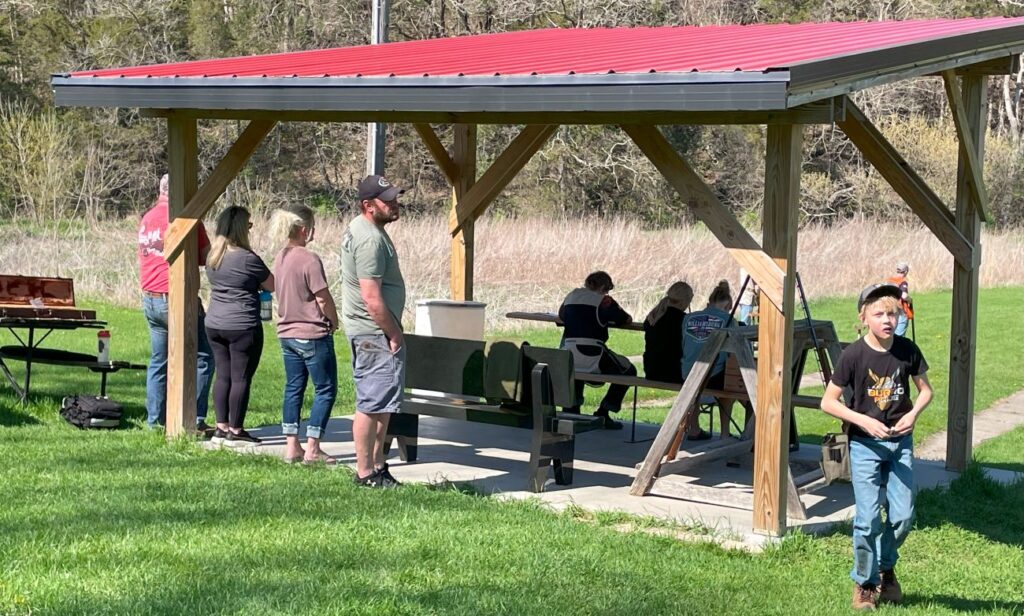 A group on people sit and stand under a red metal awning in a grassy field.