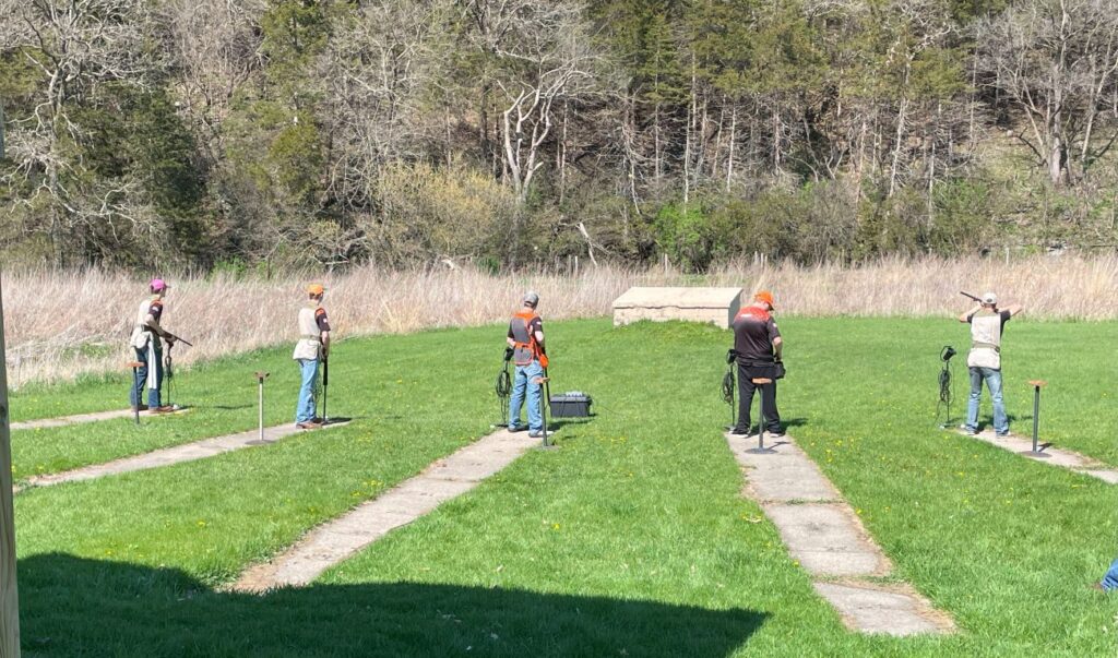 Students stand in grassy field at the ends of cement strips. The one on the right is aiming his shotgun at a clay pigeon.