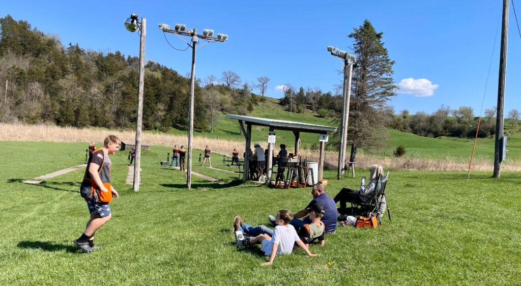 Groups of people of mixed ages sit on a grassy field and watch trap shooting practice.